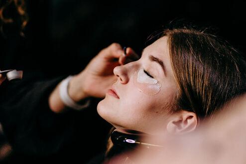 Eine Kosmetikerin trägt sorgfältig eine glitzernde Gesichtsmaske auf das Gesicht einer entspannten Kundin in einem dunklen Salon auf.