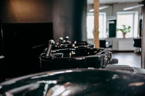 Schwarz glänzende Friseurstühle und -Waschbecken, beleuchtet durch natürliches Licht vom Fenster.