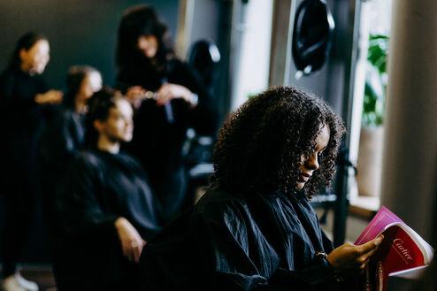 Eine Frau mit lockigem Haar liest entspannt ein Magazin, während sie in einem belebten Friseursalon auf ihre Behandlung wartet.