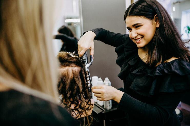 Eine fröhliche Friseurin verwendet ein Lockeneisen, um das blonde Haar einer Kundin zu stylen, die ihr im Spiegel zusieht.